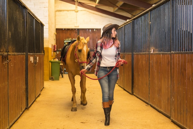 Mulher caucasiana de vaqueira passeando com um cavalo em um estábulo, chapéus do sul dos Estados Unidos, camisa xadrez rosa e jeans