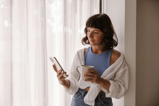 Mulher caucasiana de visão completa olhando para o telefone perto da janela