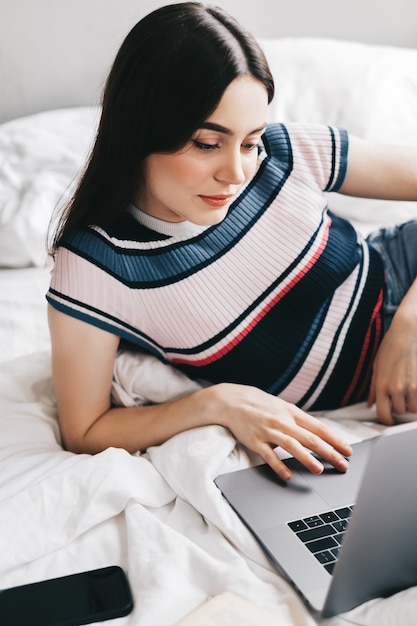 Mulher caucasiana, descansando em uma cama em casa e usando o computador portátil.