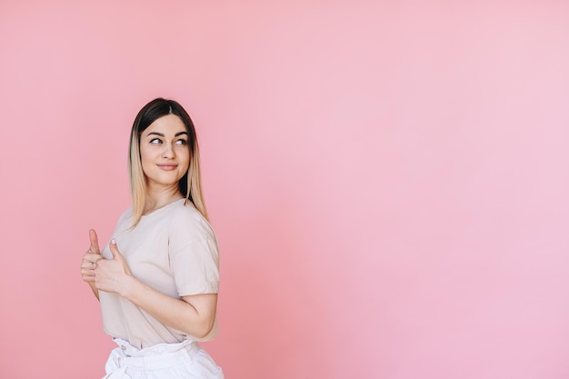 Mulher caucasiana em fundo rosa em camiseta cinza parece ao lado com as mãos levantadas que mostram os polegares