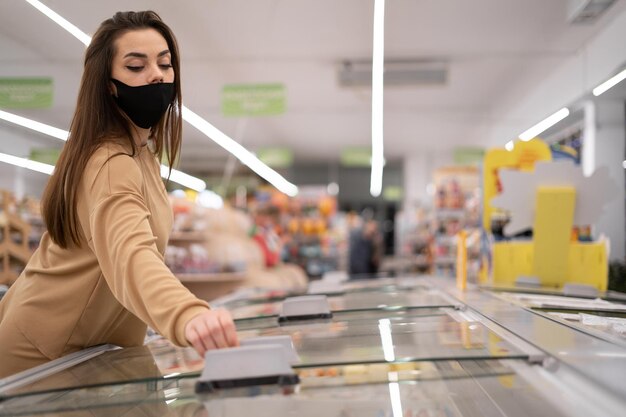 Mulher caucasiana empurrando um carrinho atravessa o supermercado Um protetor facial é usado e abre um grande freezer de exibição