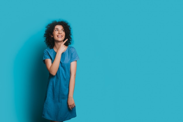 Foto mulher caucasiana encantadora com cabelo encaracolado está posando com um vestido azul e apontando para o espaço em branco perto dela