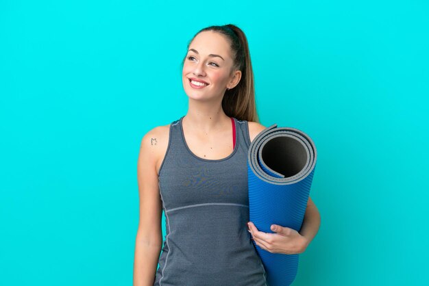 Mulher caucasiana esportiva jovem indo para aulas de ioga enquanto segura um tapete isolado em fundo azul pensando em uma ideia enquanto olha para cima