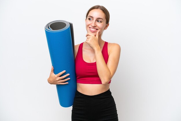 Mulher caucasiana esportiva jovem indo para aulas de ioga enquanto segura um tapete isolado no fundo branco, olhando para o lado e sorrindo