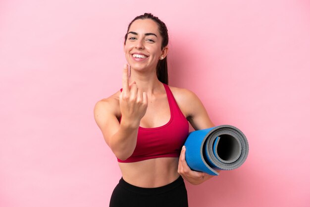 Mulher caucasiana esportiva jovem indo para aulas de ioga enquanto segura um tapete isolado no fundo rosa fazendo o gesto de chegada