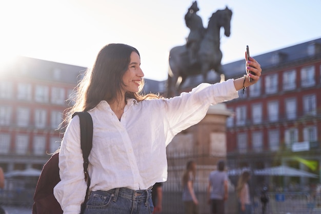 Mulher caucasiana feliz está tirando uma selfie sorrindo para a câmera em frente ao Monumento Equestre ao Rei Felipe III da Espanha na Plaza Mayor em Madrid