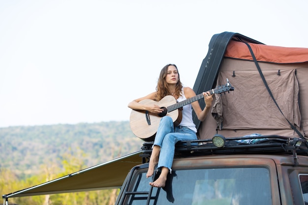 Mulher caucasiana feliz gosta de tocar violão e cantar música, belas férias no carro com a barraca no telhado. caravana em belas férias