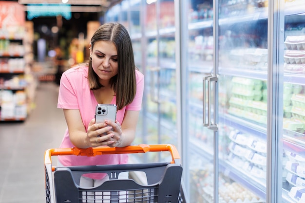 Mulher caucasiana feliz usando telefone celular em compras de supermercado Aplicação de lista de verificação comprando produtos alimentícios no supermercado