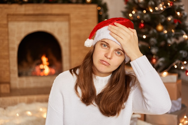 Mulher caucasiana insalubre vestindo suéter branco e chapéu de Papai Noel, sentado perto da lareira e a árvore de natal e mantendo a mão na testa, sofrendo de dor de cabeça, sintomas de gripe.