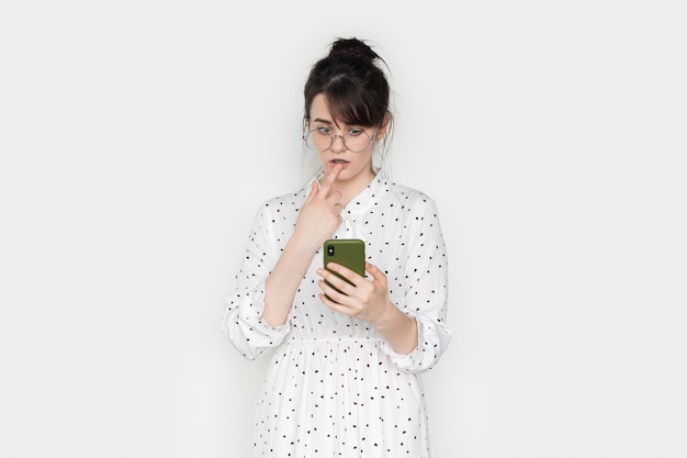 Mulher caucasiana isolada no fundo branco, olhando para a tela do telefone com expressão duvidosa e cética