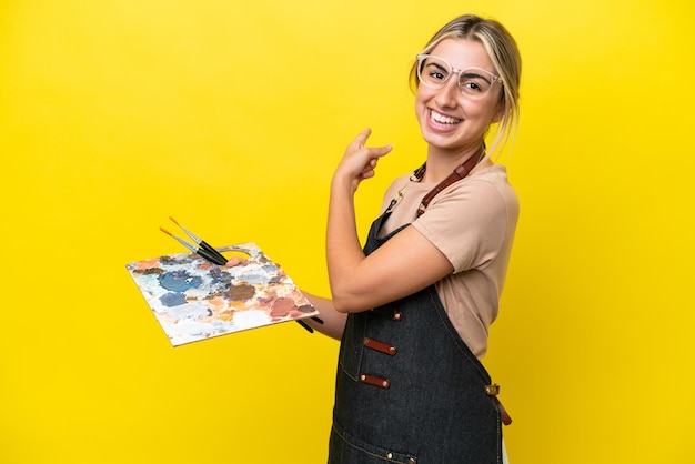 Foto mulher caucasiana jovem artista segurando uma paleta isolada em fundo amarelo, apontando para trás