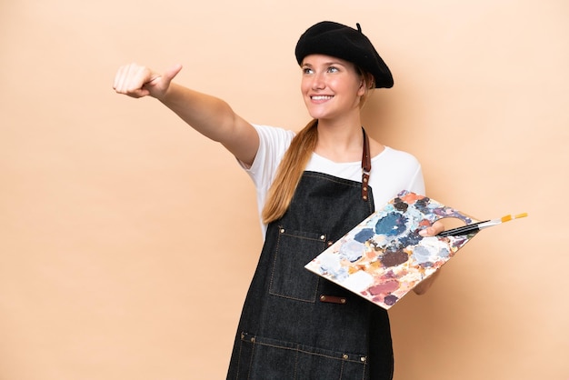 Mulher caucasiana jovem artista segurando uma paleta isolada em fundo bege, dando um polegar para cima gesto