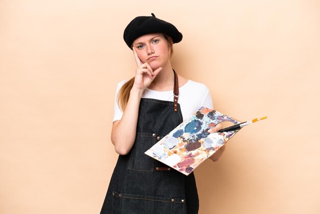 Mulher caucasiana jovem artista segurando uma paleta isolada em fundo bege, pensando em uma ideia