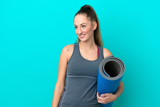 Mulher caucasiana jovem esportiva indo para aulas de ioga enquanto segura um tapete isolado em fundo azul, olhando para o lado e sorrindo