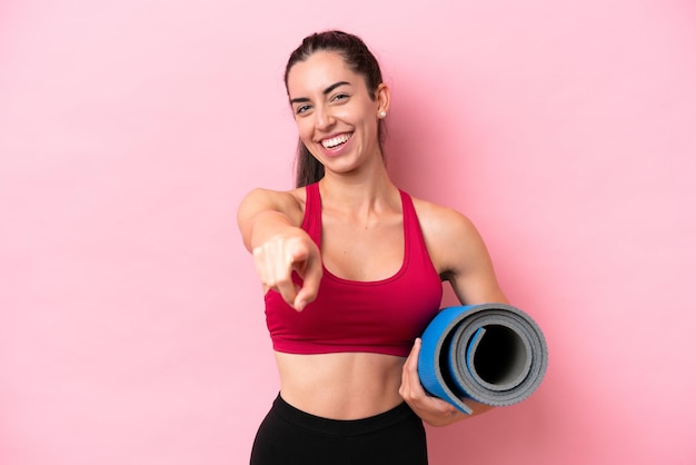 Mulher caucasiana jovem esportiva indo para aulas de ioga enquanto segura um tapete isolado no fundo rosa, apontando para a frente com expressão feliz