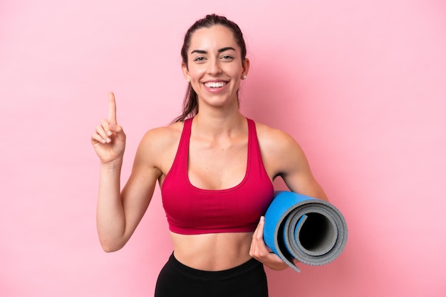 Mulher caucasiana jovem esportiva indo para aulas de ioga enquanto segura um tapete isolado no fundo rosa apontando uma ótima idéia