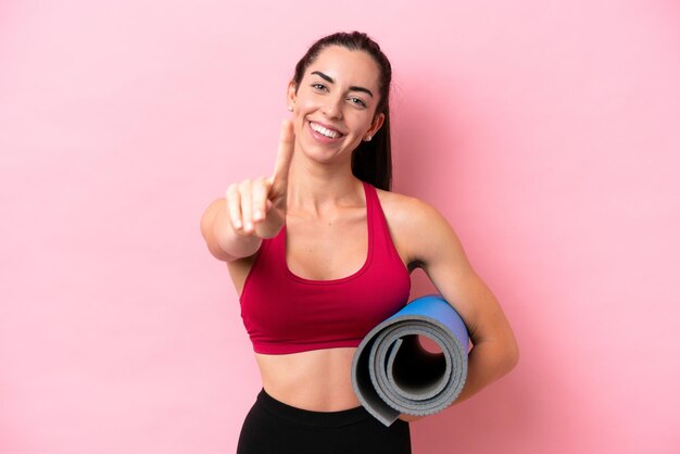 Mulher caucasiana jovem esportiva indo para aulas de ioga enquanto segura um tapete isolado no fundo rosa mostrando e levantando um dedo