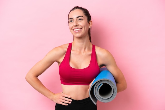 Mulher caucasiana jovem esportiva indo para aulas de ioga enquanto segura um tapete isolado no fundo rosa posando com os braços no quadril e sorrindo