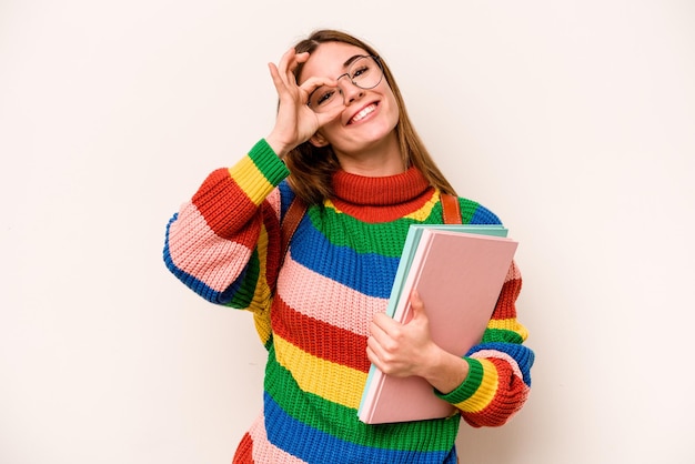 Mulher caucasiana jovem estudante isolada em fundo branco animada mantendo o gesto ok no olho