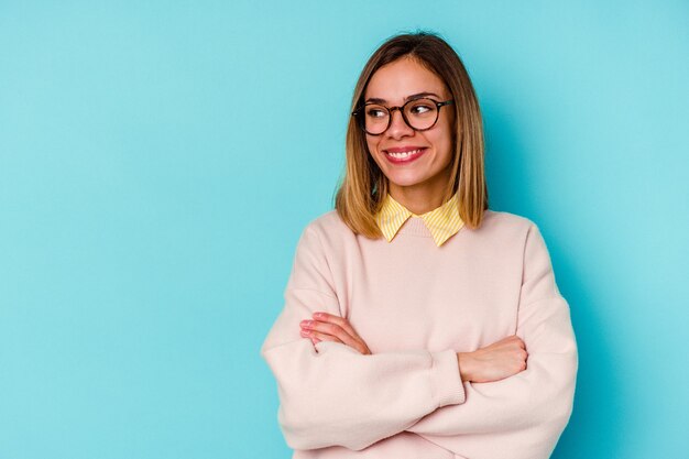 Mulher caucasiana jovem estudante isolada em um fundo azul, sorrindo confiante com os braços cruzados.