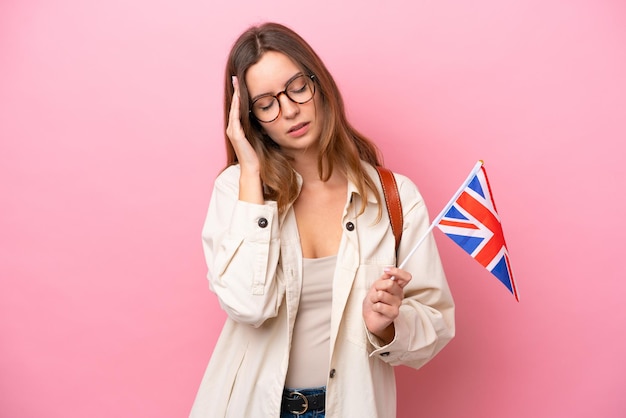 Mulher caucasiana jovem estudante segurando uma bandeira do Reino Unido isolada em fundo rosa com dor de cabeça