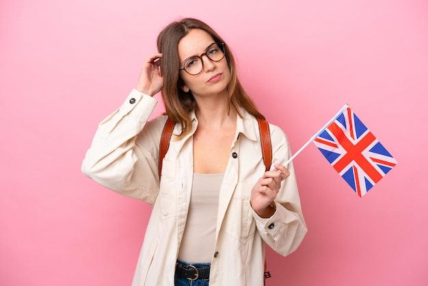 Mulher caucasiana jovem estudante segurando uma bandeira do Reino Unido isolada em fundo rosa com dúvidas