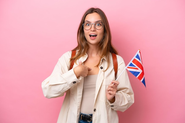 Mulher caucasiana jovem estudante segurando uma bandeira do Reino Unido isolada em fundo rosa com expressão facial de surpresa