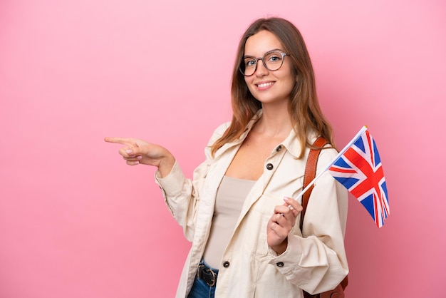 Mulher caucasiana jovem estudante segurando uma bandeira do Reino Unido isolada no fundo rosa, apontando o dedo para o lado