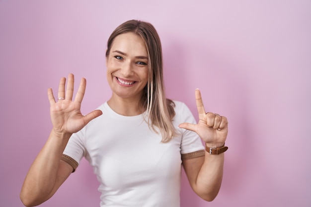 Mulher caucasiana loira em pé sobre fundo rosa mostrando e apontando para cima com os dedos número sete enquanto sorria confiante e feliz.