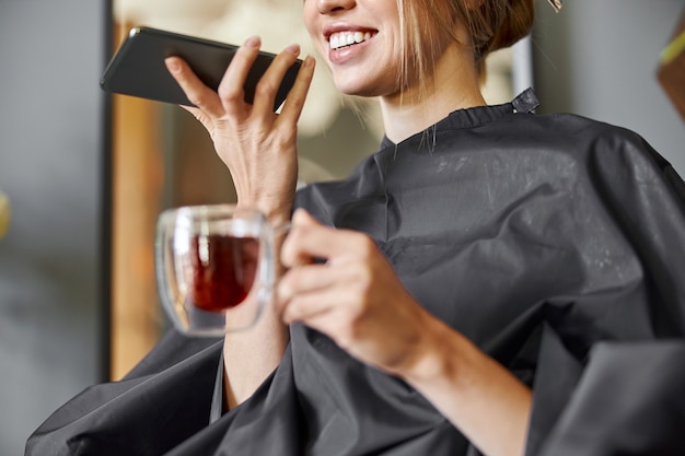 Mulher caucasiana loira feliz usando o smartphone enquanto está sentado na cadeira e esperando no salão de cabeleireiro