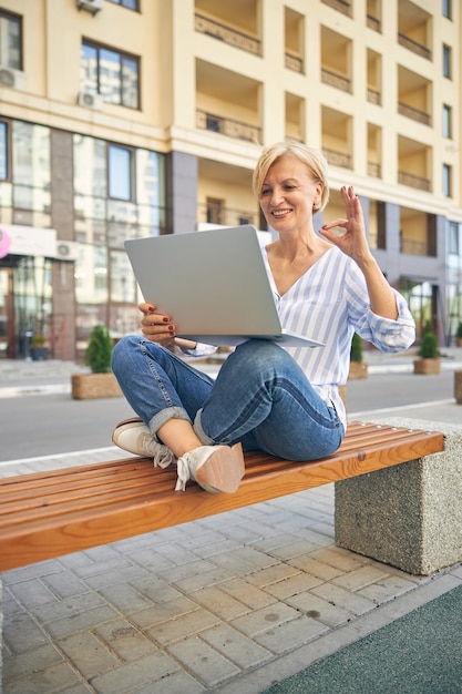 Mulher caucasiana loira madura moderna e satisfeita demonstrando um gesto de anel para seu colega via skype