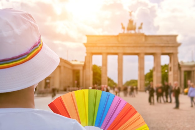 Mulher caucasiana madura LGBT em Berlim com ventilador de arco-íris e fita no chapéu branco de verão. Arco-íris, símbolo do orgulho gay LGBTQIA, diversidade. Mulher usando máscara PP2. Céu dramático sobre o Portão de Brandemburgo.