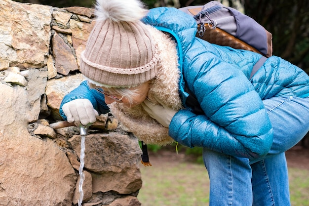 Mulher caucasiana madura sacia sua sede em uma fonte enquanto caminhava nas montanhas Mulher com roupas de inverno carrega mochila e bebe água de uma pequena fonte