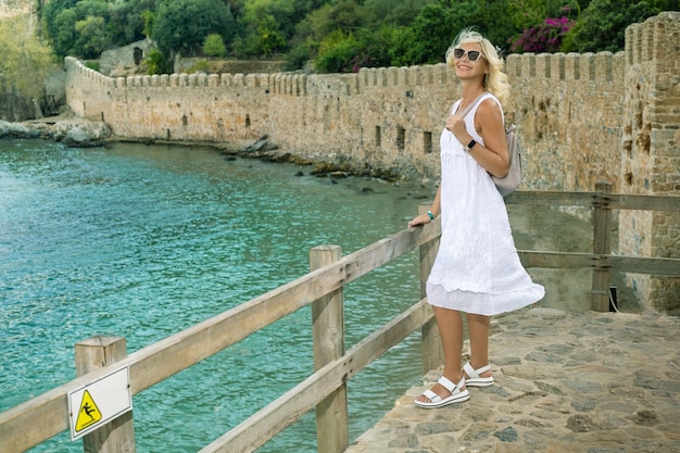 Mulher caucasiana madura sorridente em vestido branco de verão olhando para a câmera e desfrutando de uma vista da cidade de Alanya Turquia aproveita as férias de verão