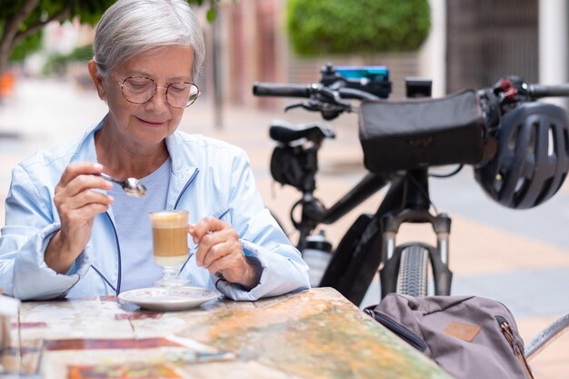 Mulher caucasiana madura sorridente, sentada em uma mesa ao ar livre, segurando um copo de café e leite nas mãos, sentada pensativamente perto de sua bicicleta elétrica