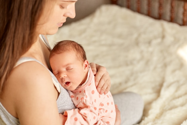 Mulher caucasiana mãe sentada na cama e segurando o bebê com sono infantil no braço, feminino tocando as costas do filho, mamãe olhando para o filho e para acalmar o bebê e dormir.