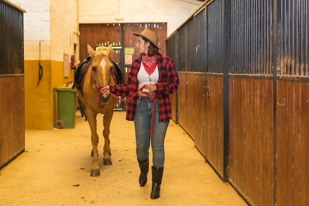 Mulher caucasiana, morena, cowgirl, passeando com um cavalo em um estábulo, usando chapéus de cowboy americanos, camisa xadrez vermelha e jeans