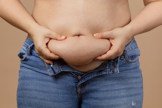 Mulher caucasiana obesa tocando e mostrando a barriga grande vestindo jeans azul descompactado em fundo bege ganho de peso repentino gordura visceral corpo positivo roupinhas apertadas necessidade de mudança de guarda-roupa