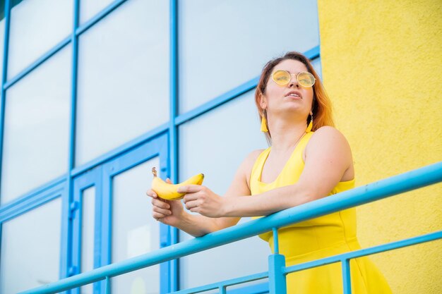 Mulher caucasiana ruiva de óculos escuros e vestido amarelo segurando uma banana ao ar livre