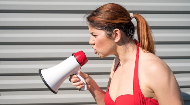 Mulher caucasiana ruiva em um biquíni vermelho gritando em um megafone contra um fundo de uma parede ondulada cinza Garota em um maiô com um alto-falante Férias de verão