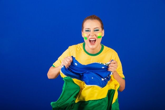 Mulher caucasiana ruiva fã de futebol do Brasil Fundo azul brasileiro gritando gol com bandeira nas mãos