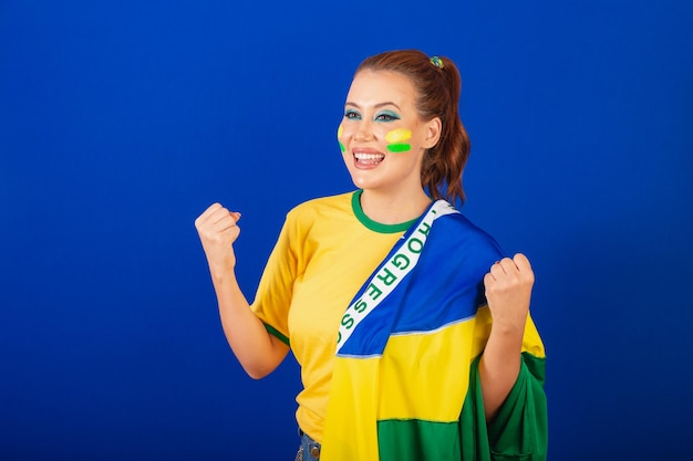 Mulher caucasiana, ruiva, fã de futebol do Brasil, fundo azul brasileiro, usando bandeira do Brasil comemorando
