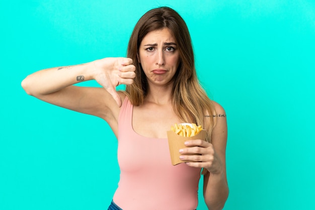 Mulher caucasiana segurando batatas fritas isoladas em um fundo azul, mostrando o polegar para baixo com expressão negativa