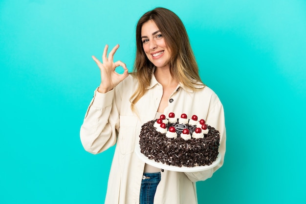 Mulher caucasiana segurando bolo de aniversário isolado em fundo azul e mostrando sinal de ok com os dedos