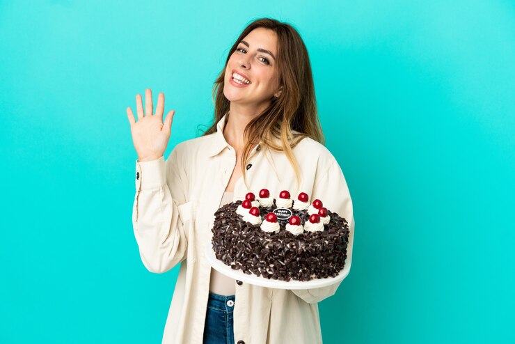 Foto mulher caucasiana segurando bolo de aniversário isolado em fundo azul e saudando com a mão com expressão feliz