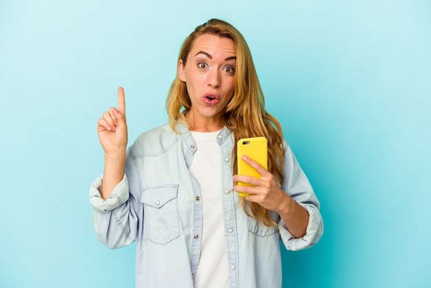 Mulher caucasiana, segurando o celular isolado em um fundo azul, tendo uma ótima ideia, o conceito de criatividade.