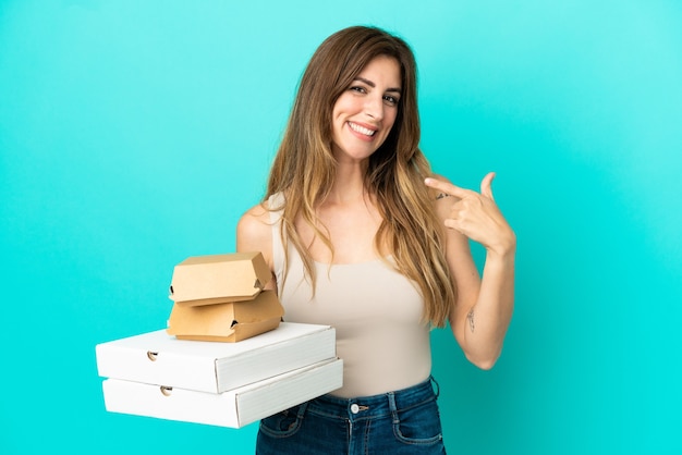 Mulher caucasiana segurando pizzas e hambúrguer isolados em um fundo azul e fazendo um gesto de polegar para cima