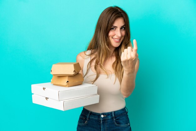 Mulher caucasiana segurando pizzas e hambúrguer isolados na parede azul fazendo um gesto próximo