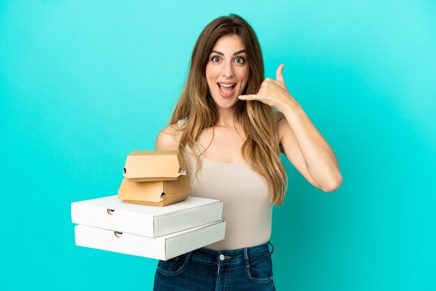 Mulher caucasiana, segurando pizzas e hambúrguer isolados sobre fundo azul, fazendo gesto de telefone. Ligue-me de volta sinal