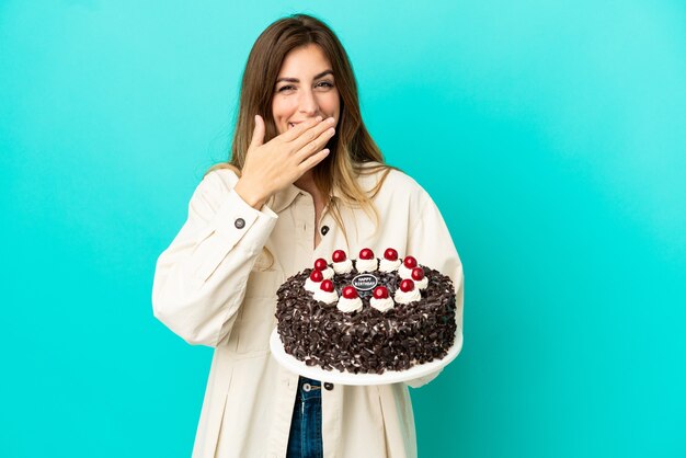 Mulher caucasiana segurando um bolo de aniversário isolado em um fundo azul, feliz e sorridente, cobrindo a boca com a mão